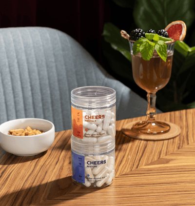 Jar of 'Cheers' protein powder on a wooden table with a cocktail and snacks in the background.