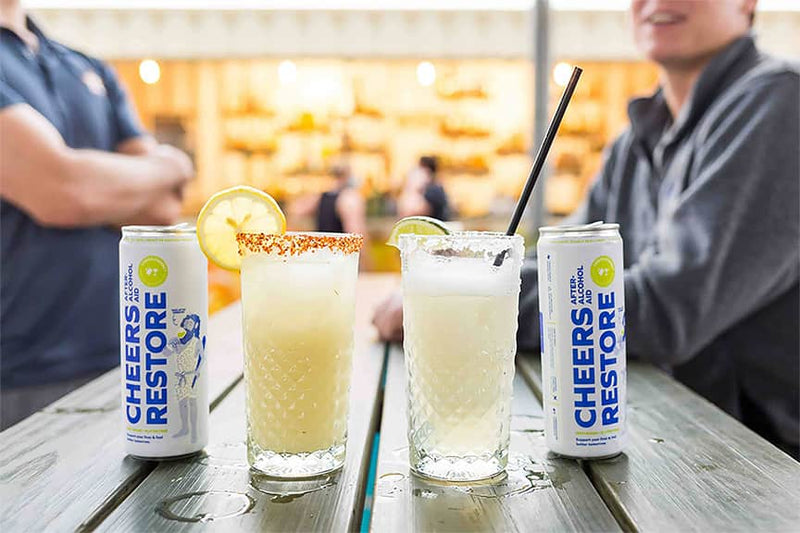 Two cans of cheers restore with two other cocktails on a bar rail