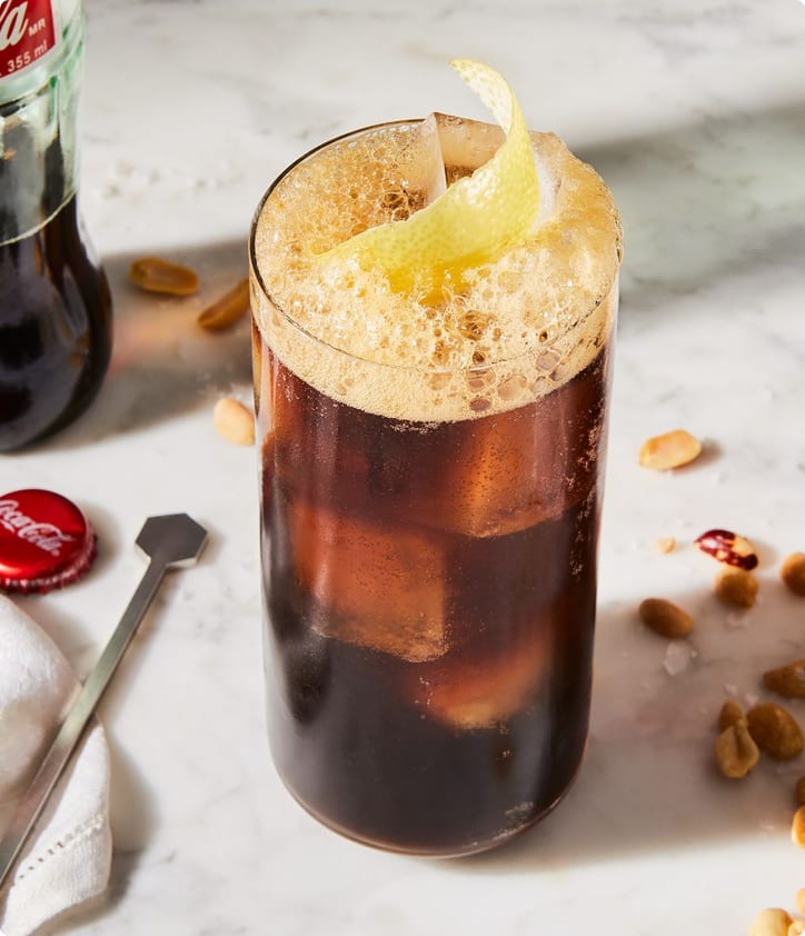 Cold brew coffee with foam and a lemon twist in a glass, with Coca-Cola bottle and ingredients on a marble surface.