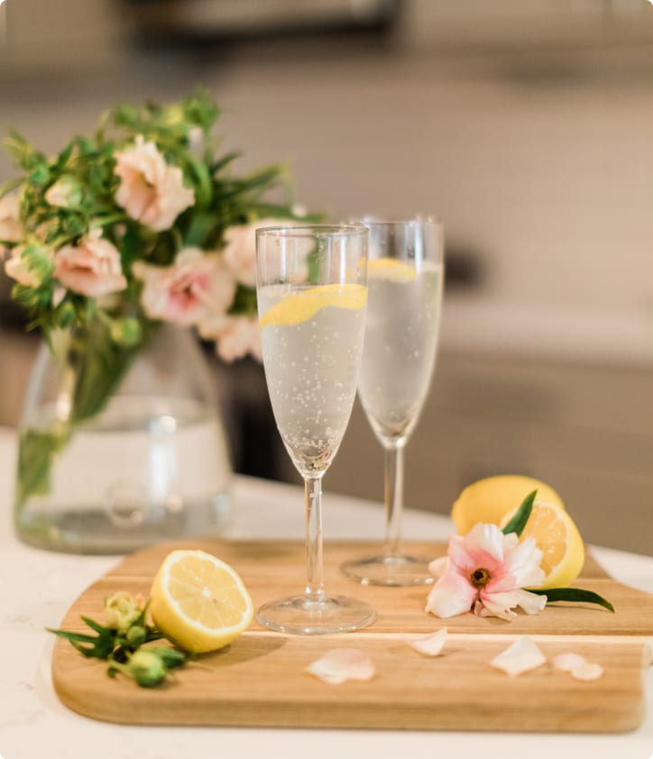 Two champagne flutes with lemon slices on a wooden board, flowers, and lemons in the background.