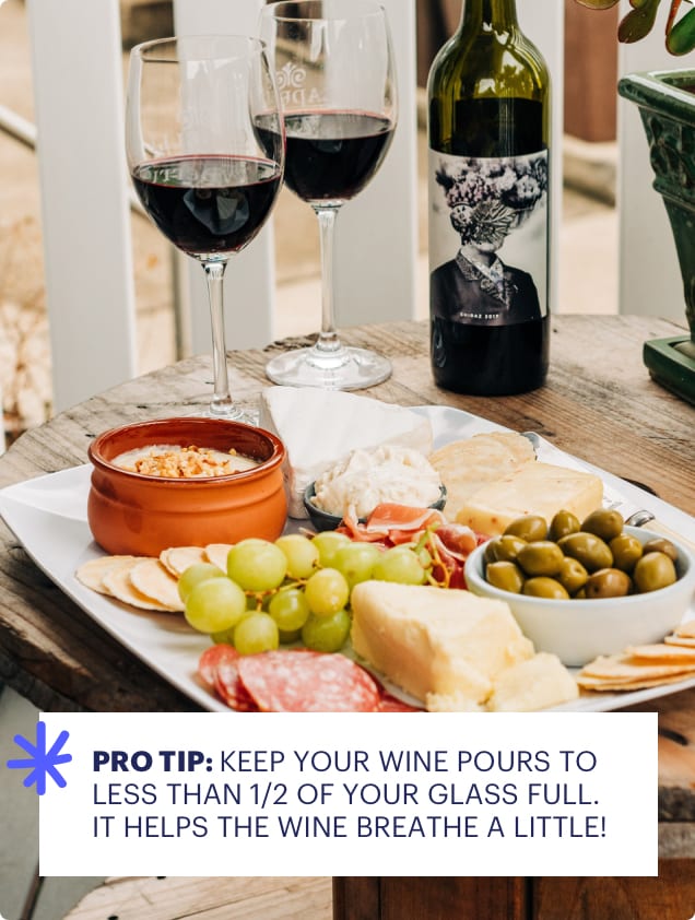 Wine bottle, glasses, and charcuterie board on a wooden table with outdoor background.