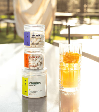 Stack of 'Cheers' supplement containers with a glass of orange juice on a table outdoors.