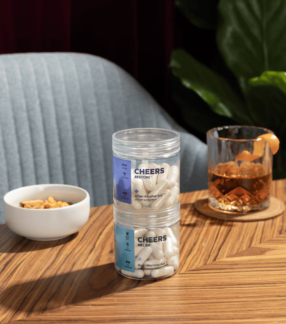 Container of 'Cheers' after-alcohol aid on a table with a glass of whiskey and snacks.