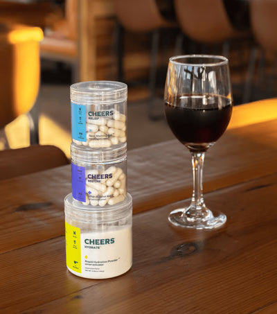 Stack of 'Cheers' supplement containers on a wooden table with a glass of dark liquid in the background.