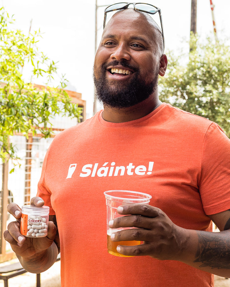 Man wearing an orange 'Sláinte!' t-shirt holding a cup and container outdoors.