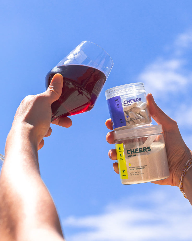 Two hands holding a glass of red wine and two containers labeled 'Cheers' against a blue sky.