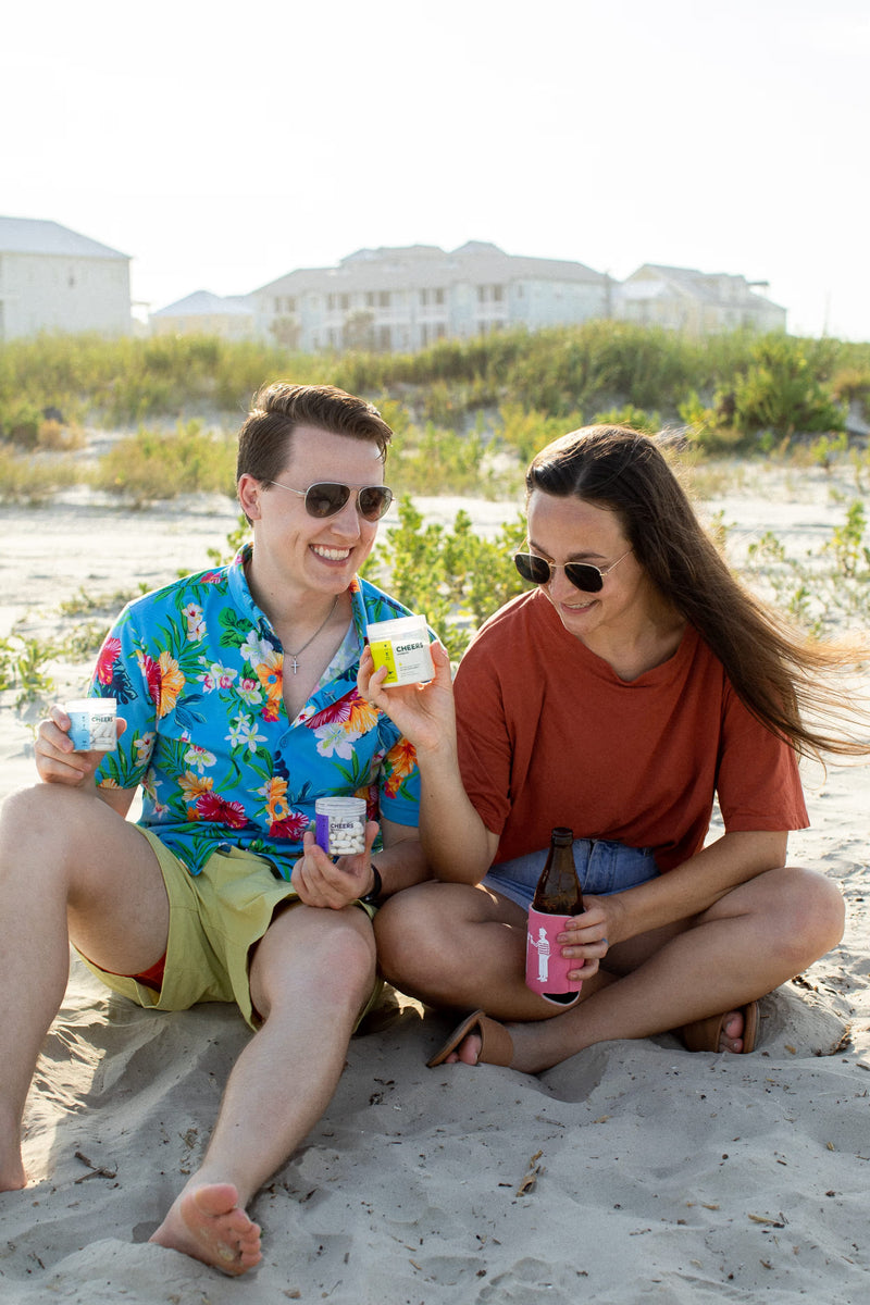 Two people sitting on a beach holding drinks and bottles of cheers products.