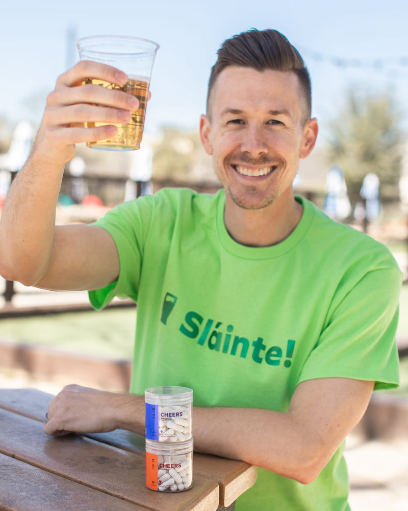 Man in a green 'Sl¡inte!' t-shirt holding a glass of beer with a bottle of cheers product on a table outdoors.