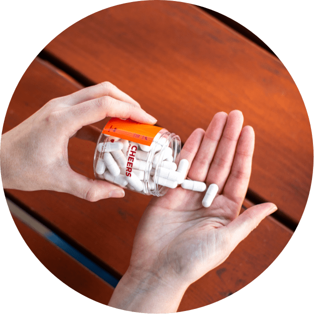 Person pouring white capsules from a bottle labeled 'Cheers' onto a palm on a wooden surface.