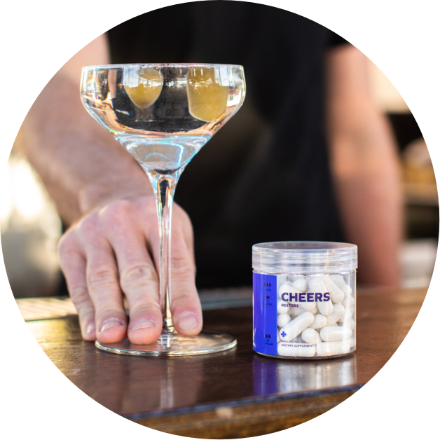 Hand reaching for a glass of wine next to a container labeled 'Cheers' on a table.