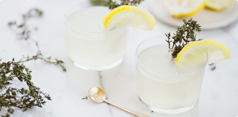Two glasses of lemonade with lemon slices and thyme on a white surface.